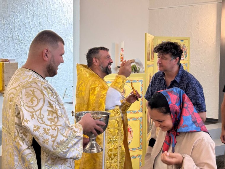 Священник кропить свяченою водою усміхнену парафіянку в хустці, поруч прислужник із посудиною